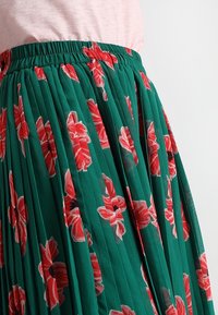 Gonna plissettata verde con un motivo floreale di fiori rossi. Vita elasticizzata, tessuto leggero e senza dettagli hardware visibili.