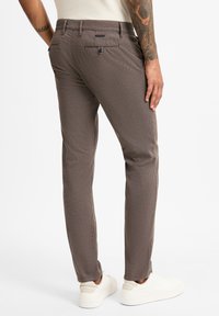 Braun gemusterte Hose mit schlanker Passform, ausgestattet mit Gesäßtaschen und einem Knopfverschluss, kombiniert mit hellen Sneakers.