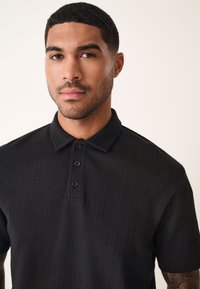 Polo noir avec un design texturé, patte de boutonnage à trois boutons et manches courtes. Présente des lignes verticales subtiles et un col standard.