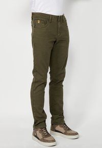 Jeans verde oliva en corte slim con bolsillos delanteros, parche trasero y detalles de costura. Combinados con zapatillas altas beige y grises.