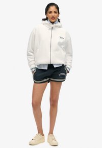 Witte fleece zip-up hoodie met een trekkoord muts, gecombineerd met zwarte shorts met witte en gele accenten. Beige sneakers maken de look compleet.