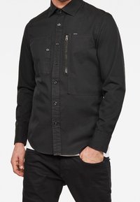 Svart långärmad skjorta i tyg, med knäppning vid kragen, två framfickor och en ficka med dragkedja på vänster bröst.
