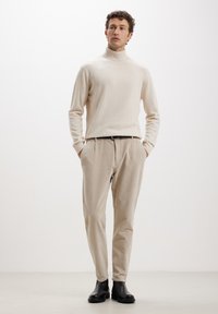 Beige Rollkragenpullover aus weichem Material, kombiniert mit hellbeigen Hosen. Schwarzer Gürtel und schwarze Stiefeletten. Schlichte, maßgeschneiderte Passform.
