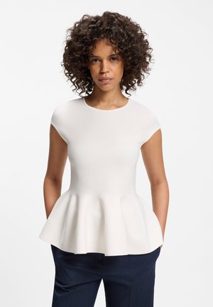 Kvinde med krøllet hår iført en hvid top med korte ærmer og peplum samt mørke bukser med tynde striber, stående mod en ensfarvet hvid baggrund.