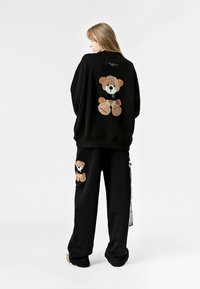 Conjunto de sudadera y pantalones negros con apliques de peluche texturizado en marrón y negro en la parte trasera y lateral. Tela suave; corte relajado.