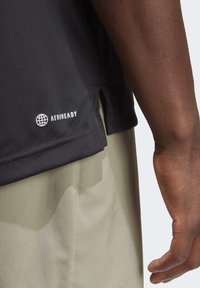 Czarna koszulka sportowa z logo AEROREADY, wykonana z gładkiego materiału, z bocznymi rozporkami. Widoczna ręka, ubrana w jasnoszare szorty.