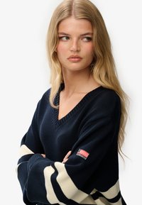 Granatowy sweter z dzianiny ma dekolt w kształcie litery V, paski w kolorze kremowym oraz małą naszywkę z logo na rękawie. Teksturowany materiał o luźnym kroju.
