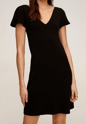 Vrouw die een korte zwarte jurk draagt met vleermuismouwen en een V-hals, staand tegen een effen lichte achtergrond.