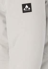Helles graues Jackett mit glatter Textur. Verfügt über ein schwarzes Stoffetikett mit weißem Berglogo und "WHISTLER"-Markenname auf dem oberen Ärmel.