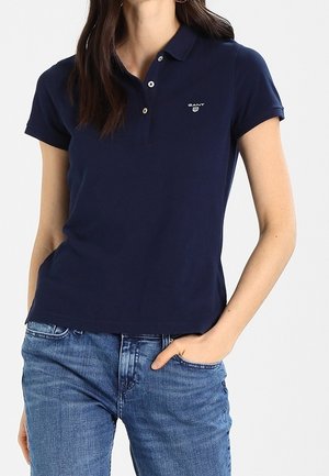 Femme portant un polo bleu marine à manches courtes avec un petit logo sur la poitrine gauche et un jean bleu en denim, main dans la poche.