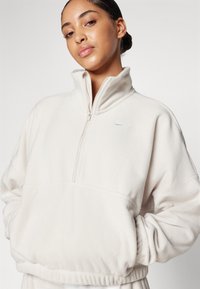 Light beige fleece pullover with a high collar, half-zip opening, and ribbed hem. Features a small, contrasting logo on the chest.