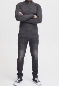 Grauer Reißverschluss-Pullover mit gerippten Bündchen und Saum, kombiniert mit dunkler Jeans und schwarzen Sneakers mit weißen Sohlen und Schnürsenkeln.