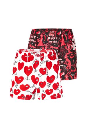 Shirtless man with curly hair and beard wearing white boxer shorts patterned with red cartoon hearts on a plain background.
