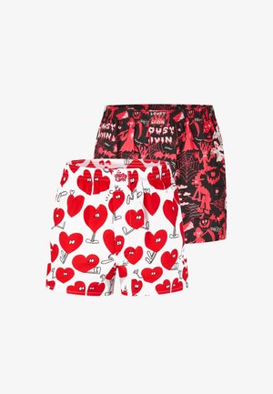 Oberkörperfreier Mann mit lockigem Haar und Bart, der weiße Boxershorts mit roten Cartoon-Herzen auf einfarbigem Hintergrund trägt.