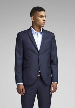 Completo blu navy con un design aderente, giacca monopetto e rever a punta. Camicia azzurro chiaro sotto la giacca; dettagli di bottoni e tasche visibili.