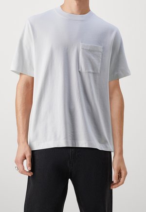 Man wearing plain white short-sleeve t-shirt with chest pocket and black pants against plain background.