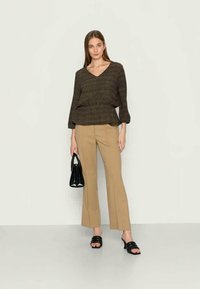 Mulher vestindo uma blusa preta com padrão, calças largas bege, sandálias pretas de salto, segurando uma bolsa preta, de pé contra um fundo liso.