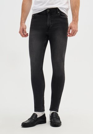 Uomo che indossa jeans neri aderenti, calzini bianchi, mocassini neri e una camicia bianca, in piedi davanti a uno sfondo chiaro uniforme.