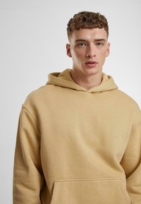 Felpa cappuccio beige realizzata in morbido tessuto, con una grande tasca frontale e una vestibilità comoda. Design minimalista senza motivi visibili.