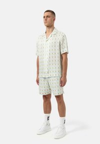 Herenkorte mouw, collared shirt en bijpassende shorts in lichtgroen met een repetitief bloemenpatroon, gecombineerd met witte sneakers en sokken.