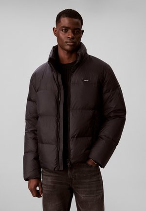 Homme portant une doudoune noire par-dessus un pull noir et un jean foncé, debout devant un fond gris uni.