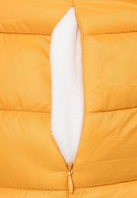 Sárga puffjacket, texturált felülettel, zipzáras zsebbel, amelyet fehér flíz bélés borít a melegség és kényelem érdekében.