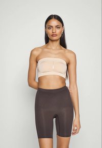 Kvinna med långt mörkt hår iklädd en strapless beige bandeau-topp och högmidjade mörkbruna cykelbyxor, stående mot en enfärgad bakgrund.