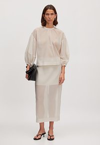 Doorzichtige, lichtbeige top met geplooide mouwen, gecombineerd met een bijpassende rok. Houdt een zwarte clutch vast, geaccentueerd door zwarte sandaaltjes met banden.