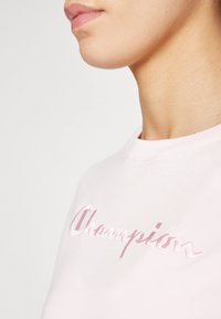 Lys rosa bomull t-skjorte med brodert "Champion" logo i en mørkere rosa, rund hals og korte ermer med en jevn tekstur.