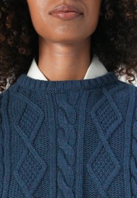 Navyblauer Strickpullover mit Kabel- und Rautenmuster, in runder Form mit strukturiertem Finish, über einem weißen Kragen getragen.