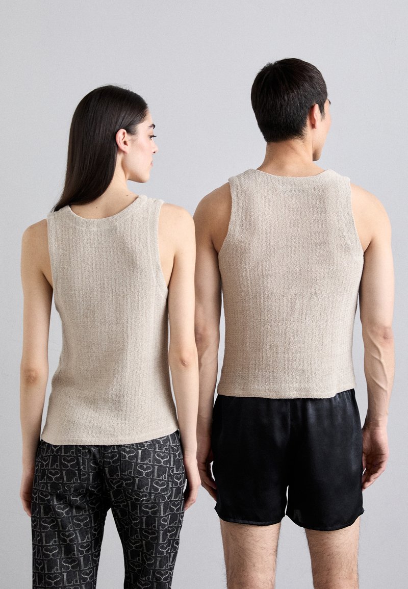 Beige, strukturierte, gestrickte ärmellose Tops, getragen von zwei Personen, wobei eine mit gemusterten Hosen kombiniert ist und die andere mit schwarzen Shorts.