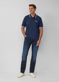 Marineblauwe polo met witte kraag en streep op de mouwen, gecombineerd met donkere denim jeans en witte schoenen, model staat met handen in de zakken.