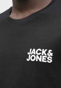 Melnas T-krekls ar baltu "JACK & JONES" logo, kas iespiests kreisajā krūtīs, valkāts personas, kas redzama no kakla līdz viduskrūtīm.