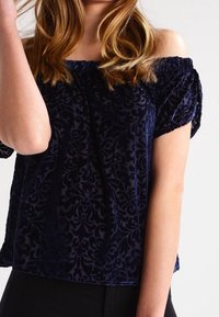 Granatowa bluzka typu off-shoulder z teksturowego aksamitu, ozdobiona kwiatowym wzorem, z krótkimi rękawami i luźnym krojem.