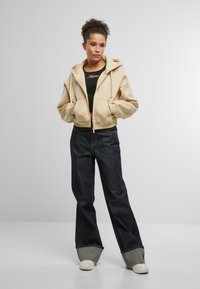 Zkrátená béžová mikina s předními kapsami, spárovaná s tmavými denimovými širokými kalhotami a bílými teniskami. Oblečení obsahuje upnutý černý top pod. 