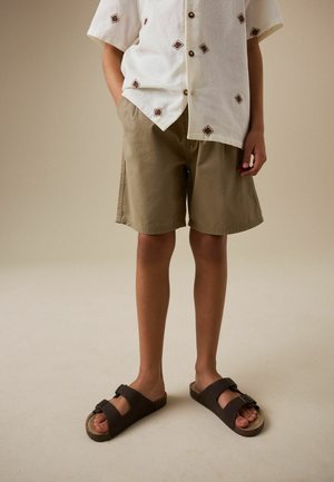 Barn iført beige knælange shorts, hvid kortærmet skjorte med brune diamantmønstre og mørkebrune sandaler, står på en neutral baggrund.