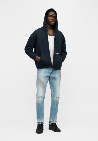 Νεαρός άνδρας στέκεται με τα χέρια στις τσέπες, φοράει μαύρο φούτερ με φερμουάρ, λευκή φανέλα, σκισμένο ανοιχτό μπλε τζιν και μαύρα αθλητικά παπούτσια.