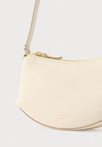 Τσάντα χειρός από κρεμ δέρμα με κυρτό σχέδιο, χρυστό φερμουάρ και λογότυπο που είναι ανάγλυφο στο κάτω μέρος. Διαθέτει λεπτό, ρυθμιζόμενο λουράκι.