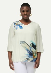 Lys creme bluse med V-hals og trekvartærmer. Har blå blomstertryk med detaljerede kronblade og blød tekstur.