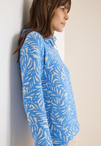 Pull-over tricoté bleu avec motif abstrait de feuilles beige, manches longues et col. Texture douce et coupe décontractée.