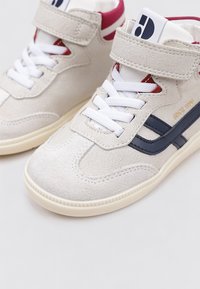Zapatillas altas de ante beige con cordones blancos, rayas en azul marino y cierre de velcro. Suela de goma crema gruesa. Detalle interior en rojo.