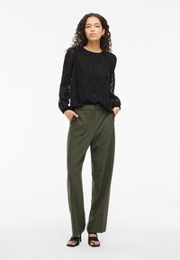 Black long-sleeve blouse with textured leaf pattern, paired with high-waisted olive green trousers. Black heeled sandals complete the outfit.