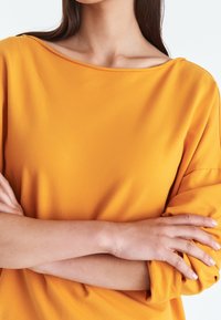 Värikäs oranssi pitkähihaisen, venepääntiepaidan kangas on sileää ja pehmeää. Paita on väljää mallia, ja hihansuut on käännetyt.