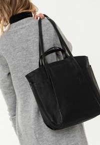 Schwarze Leder-Totetasche mit zwei Griffen, ausgestattet mit einer glatten Textur, Seitentaschen und eingeprägtem Branding. Getragen über einem grauen Pullover.
