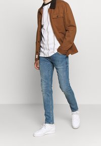 Brown jacket with a corduroy collar, white t-shirt featuring vertical stripes, blue jeans, and white sneakers. Casual outfit with layered textures.