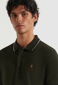 Polo shirt verde scuro con vestibilità slim, dotato di una chiusura a tre bottoni, righe bianche e bordeaux sul colletto e un logo ricamato.