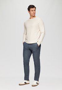 Beige strukturierte Langarmbluse, dunkelblaue karierte Hose und weiße Sneakers mit braunen Akzenten, getragen von einem stehenden männlichen Modell.