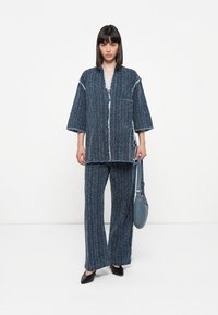 Denim two-piece outfit featuring a loose-fit top with a pocket, frayed edges, and wide leg pants in textured blue fabric, paired with a gray bag.