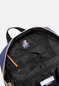 Interno di uno zaino blu con fodera nera, tasca con zip e cinghie organizzative. Presenta un'etichetta con logo e una cinghia decorativa colorata.