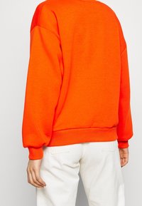 Oranžový svetr s dlouhými balónovými rukávy a žebrovanými manžetami, spárovaný s bílými kalhotami. Textura vypadá měkce a neformálně.